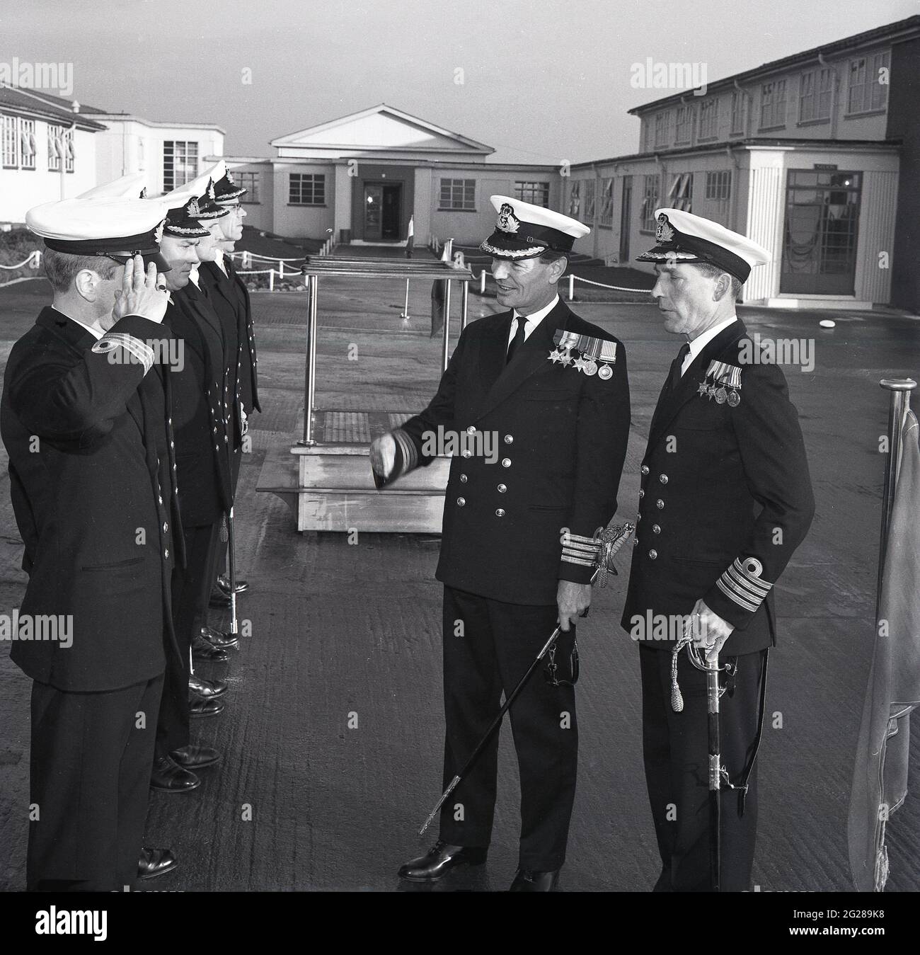 1960er Jahre, historisch, außerhalb der HMS Caledonia, in einer BefehlsüberÜbergabe, zwei Royal Naval Captains, einer, der einer Reihe von niederrangigen Offizieren, Fife, Schottland, vorgestellt wurde. Captain (Capt) ist ein hochrangiger Offizier der Royal Navy, über einem Commander und unter Commodore und hat einen NATO-Ranking-Code von OF-5. Der Rang entspricht einem Oberst der britischen Armee und der Royal Marines sowie einem Gruppenkapitän der Royal Air Force. Die HMS Caledonia wurde 1937 eröffnet und war bis 1985 für die Ausbildung von Handwerkern verantwortlich, wobei viele Tausende von jungen Männern eine Ausbildung durchgemacht haben. Stockfoto