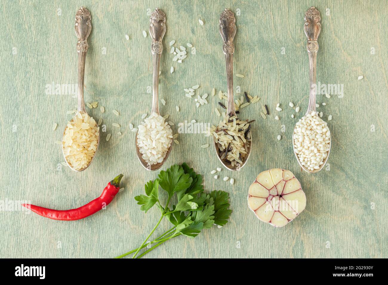 Verschiedene Reissorten in Löffeln mit Gewürzen. Wildreis, Sushi-Reis, geschredderter und gedämpfter Reis auf grünem Holzgrund mit Knoblauch, Chili und Pars Stockfoto