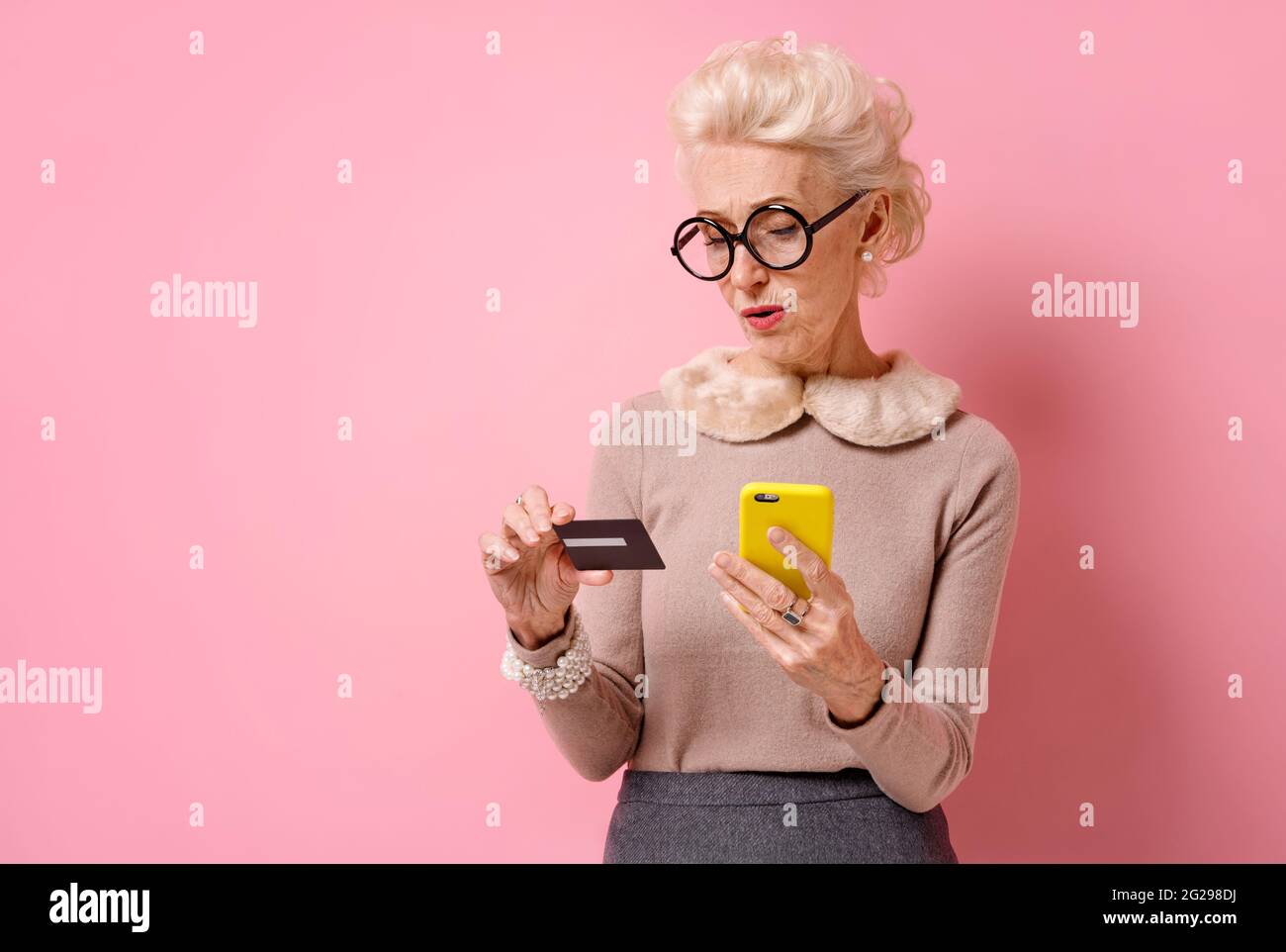 Problem mit einer Kreditkarte. Großmutter bezahlt mit Kreditkarte und Smartphone. Foto von gutherzigen älteren Frau auf rosa Hintergrund. Stockfoto