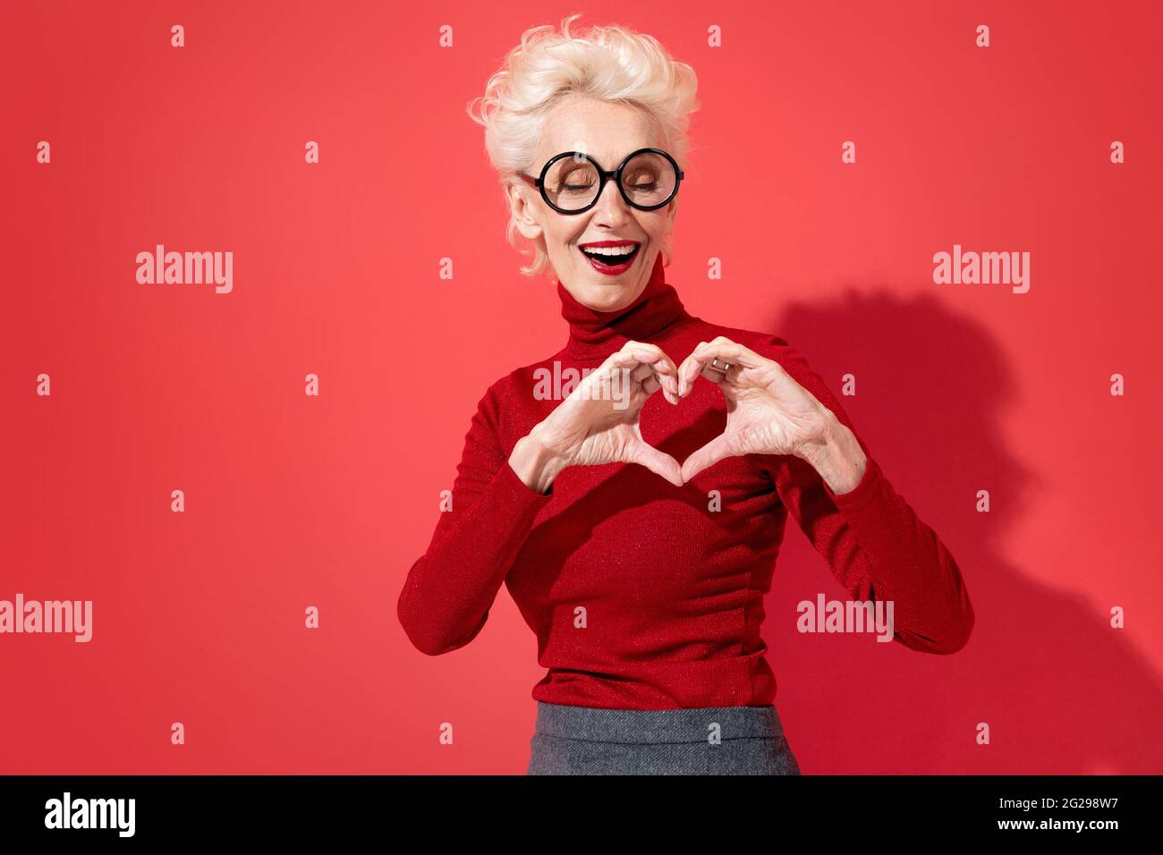 Schöne Frau zeigt Herz-Symbol, Formen Liebe Zeichen mit Händen. Foto von lächelnder älterer Frau in der Liebe auf rotem Hintergrund. Sei mein Valentinstag Stockfoto