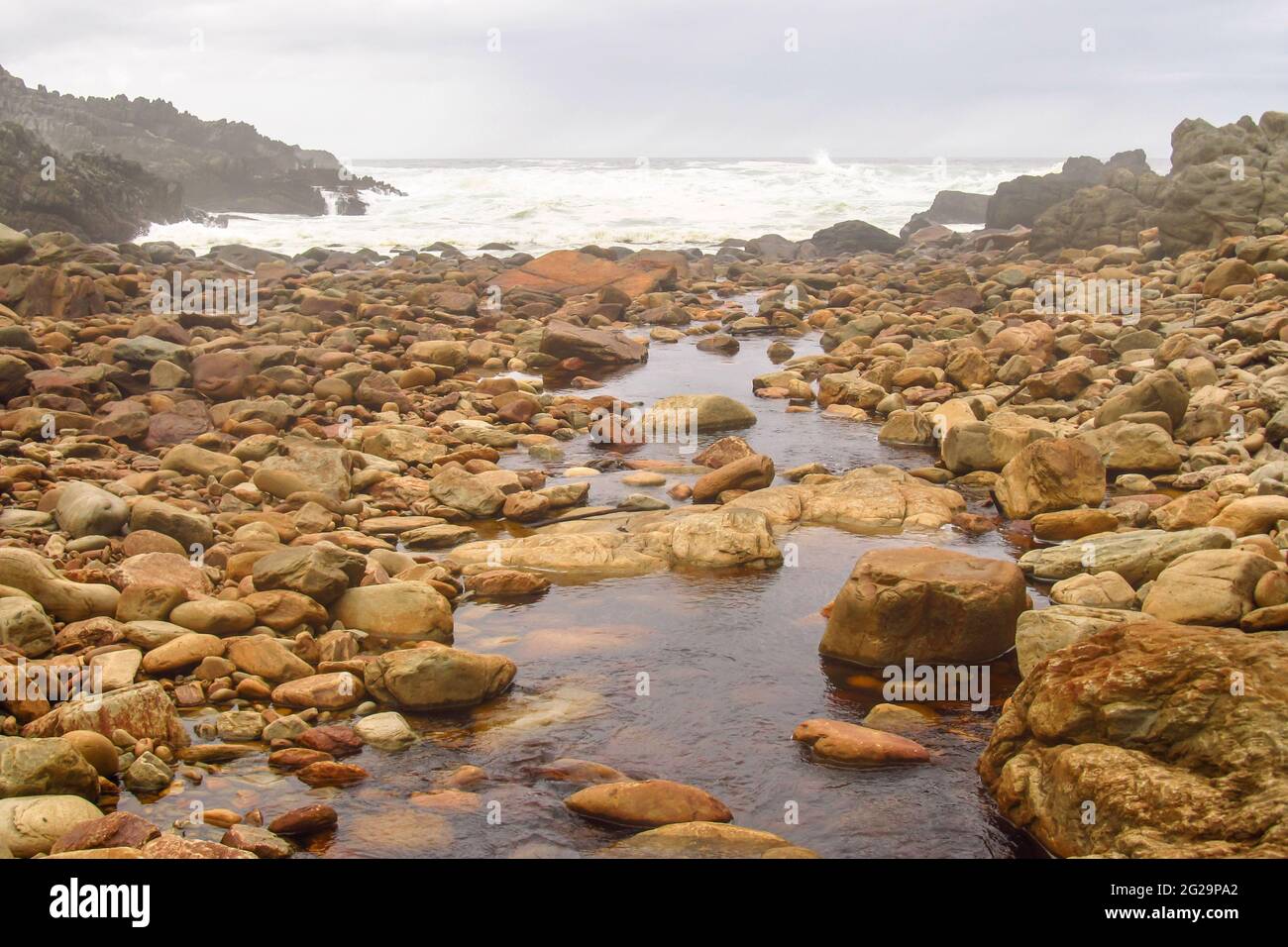 Ein kleiner Bach, der den Indischen Ozean verbindet und durch einen mit Felsbrocken bedeckten Strand entlang der Tsitsikamma Küste fließt Stockfoto