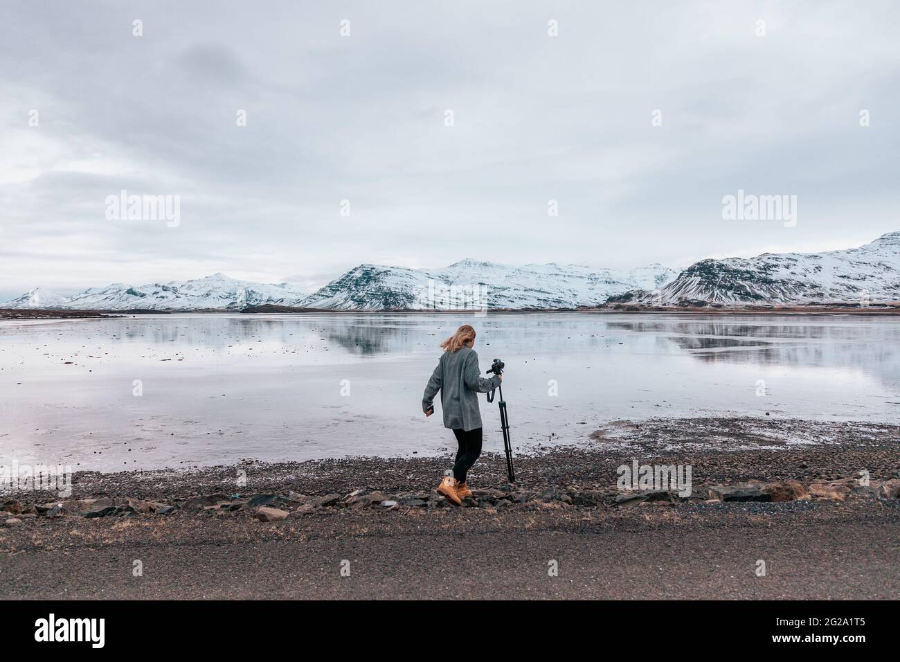 Rückansicht einer Frau, die eine professionelle Fotokamera mit Stativ trägt, die auf einer felsigen Küste des Sees im verschneiten Island unterwegs ist Stockfoto