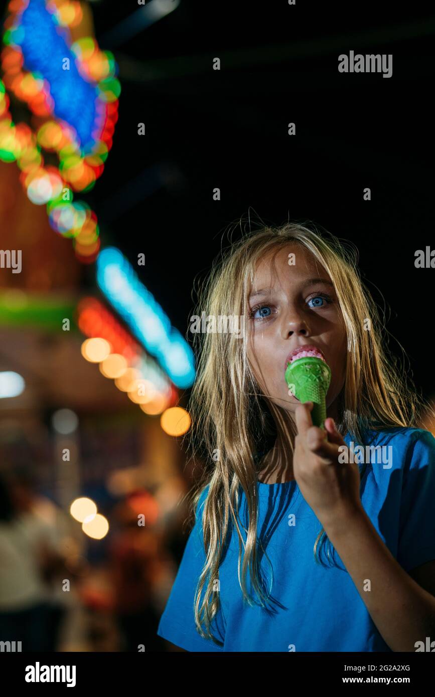 Kleines blondes Mädchen, das nachts im Vergnügungspark ein leckeres Eis isst Stockfoto