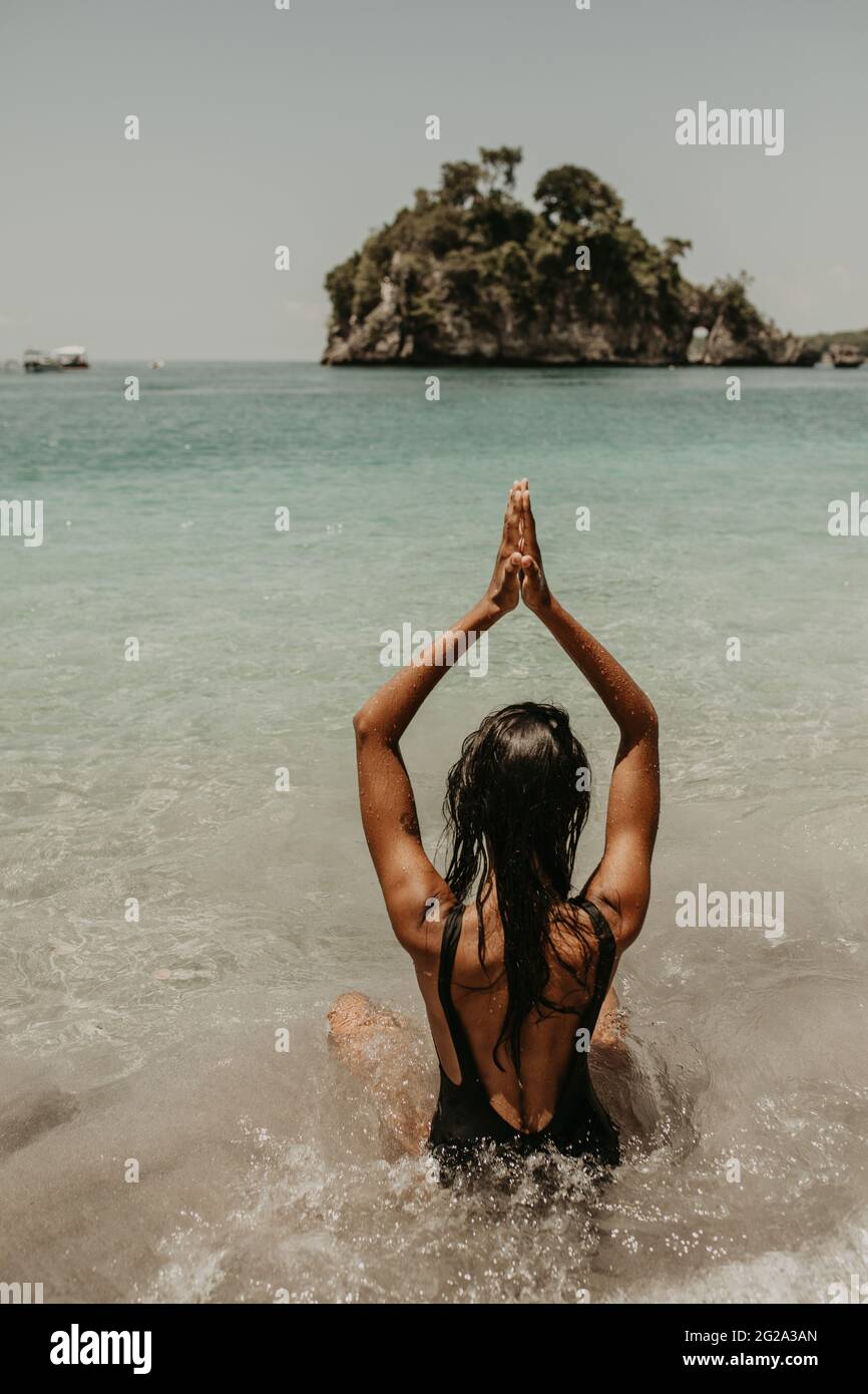 Rückansicht einer anonymen ethnischen Frau in schwarzem Bikini, die Yoga-Pose macht und allein am Meer sitzt und meditiert Stockfoto