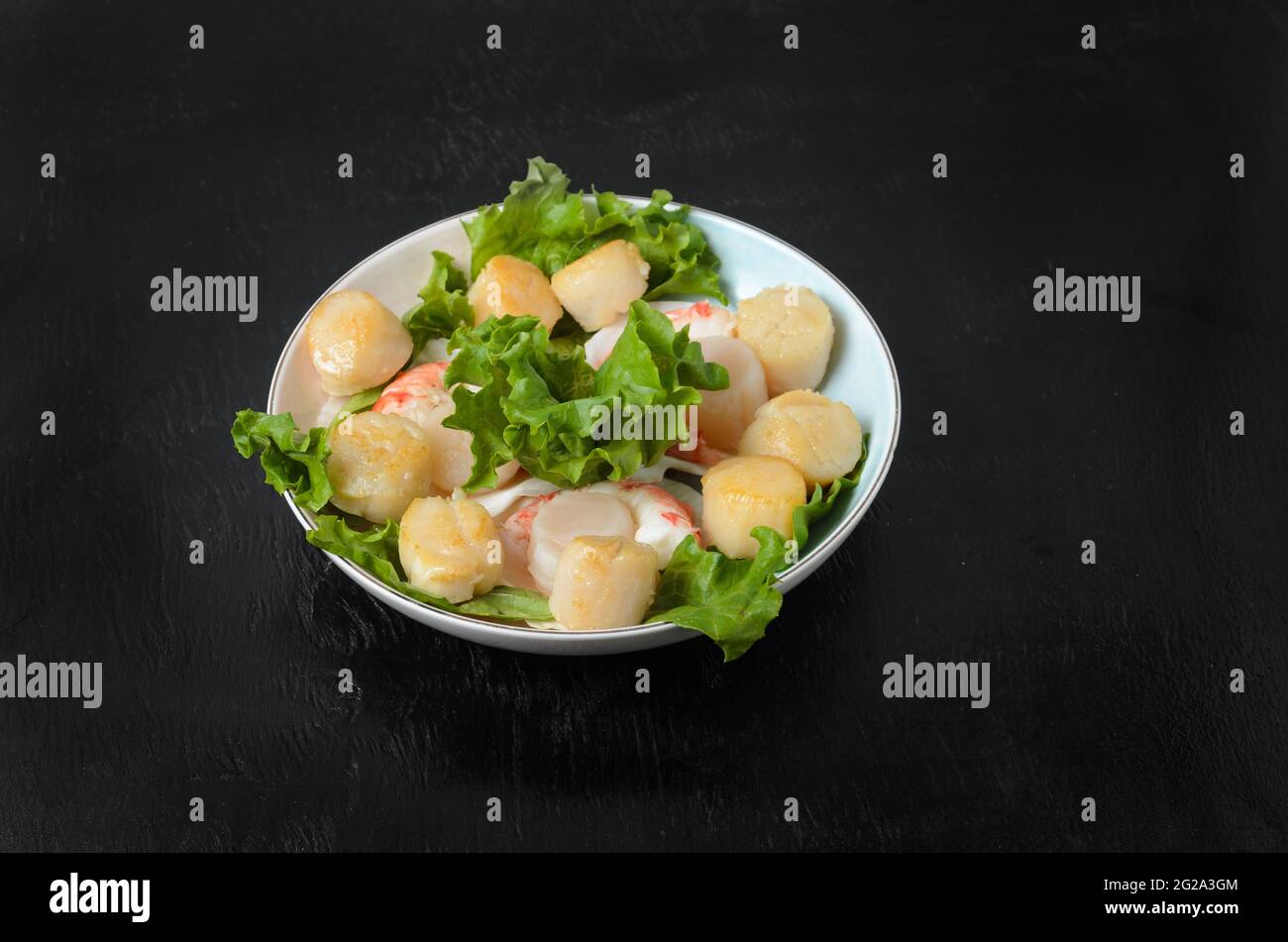 Gebratener Jakobsmuschel mit gekochtem Garnelenschwanz mit Salatblättern in einer Keramikplatte auf dunklem Hintergrund. Selektiver Fokus. Stockfoto