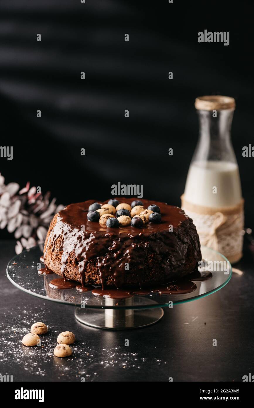 Appetitlich hausgemachter Kuchen mit Schokolade Ganache mit Nüssen und Heidelbeeren auf dem Tisch mit einer Flasche Milch auf schwarzem Hintergrund dekoriert Stockfoto
