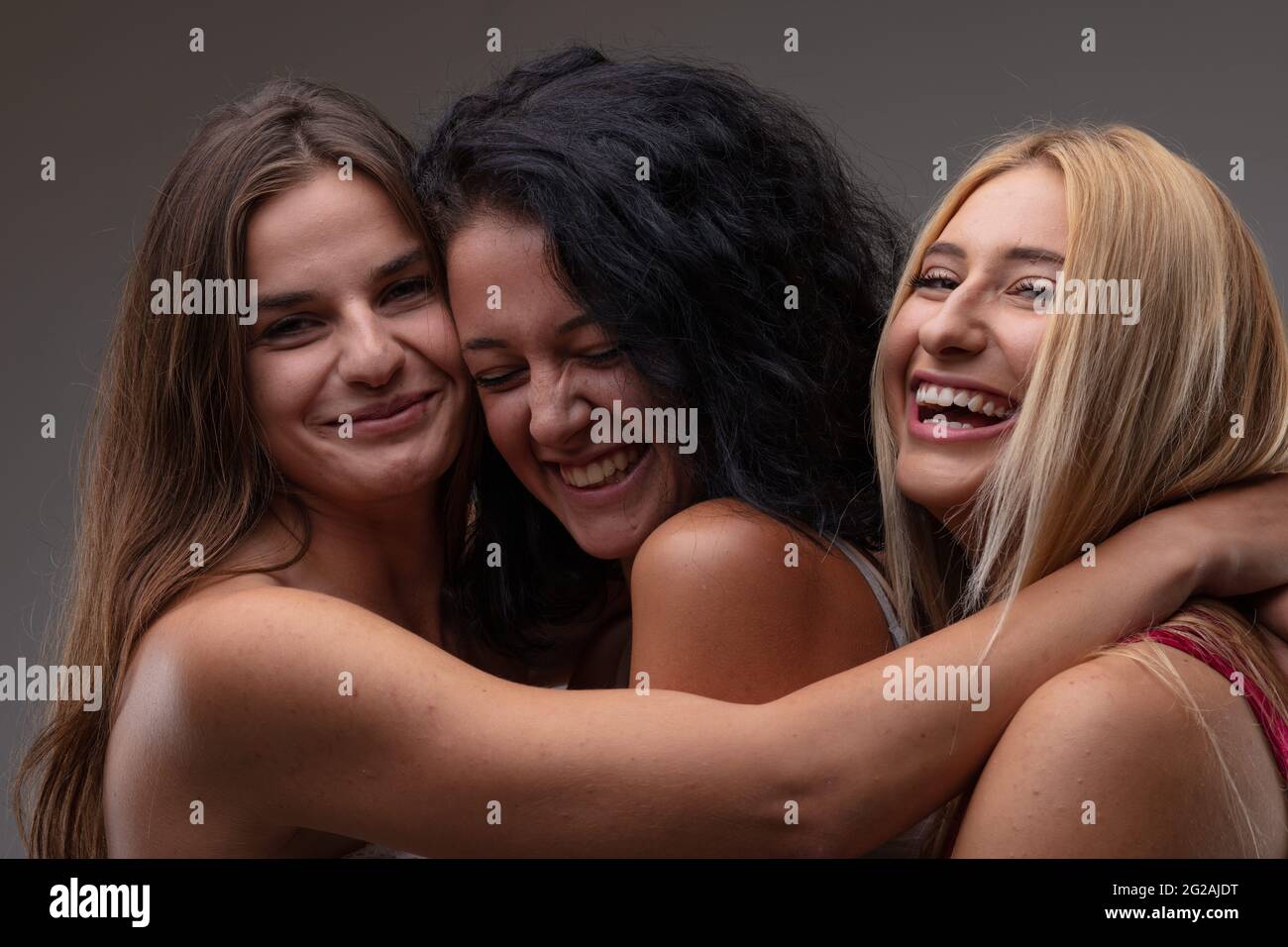 Drei sorglose, glückliche junge Frau, die über die Kamera lacht, während sie Arm in Arm in enger Umarmung in einem Kopf-Schultern-Porträt gegen einen grauen Stu posieren Stockfoto