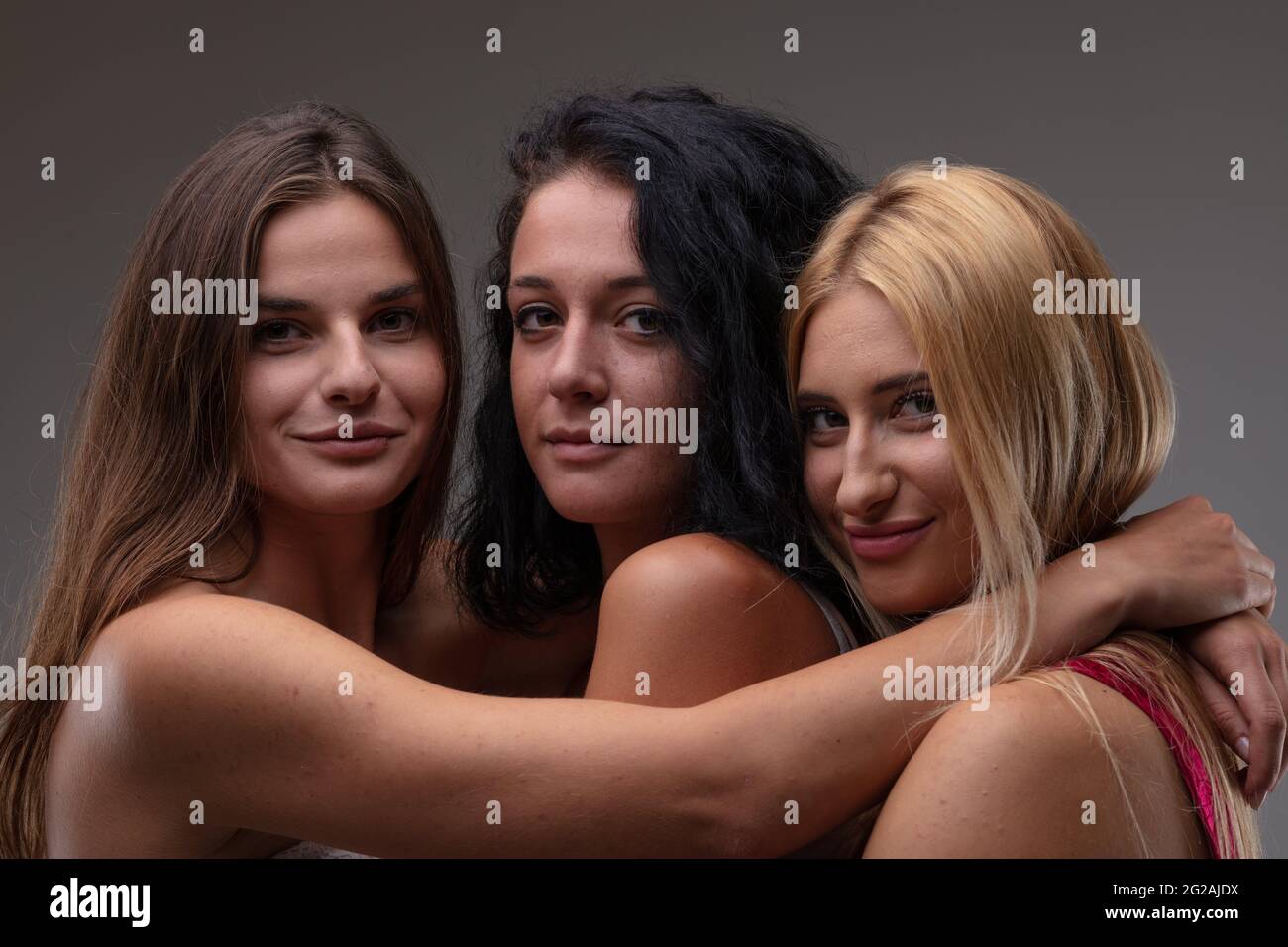 Drei attraktive, freundliche junge Frauen posieren in enger Umarmung und lächeln in einem Kopf- und Schulterporträt vor einem grauen Studio-Backgroun vor der Kamera Stockfoto