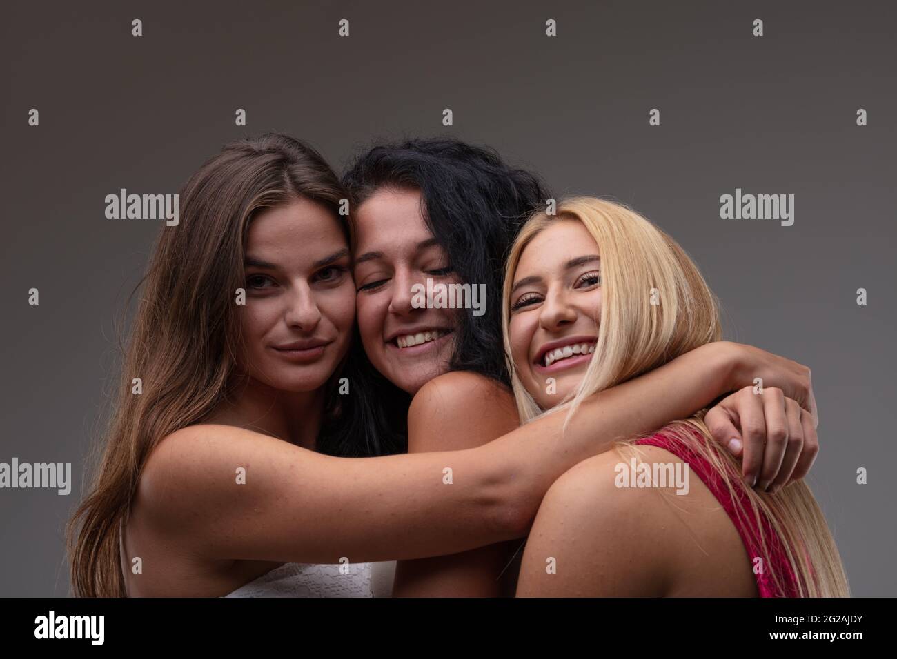 Drei glücklich lächelnde attraktive junge Frau, Arm in Arm, posiert mit ihren Köpfen dicht zusammen und grinst vor grauem Studiohintergrund vor der Kamera Stockfoto
