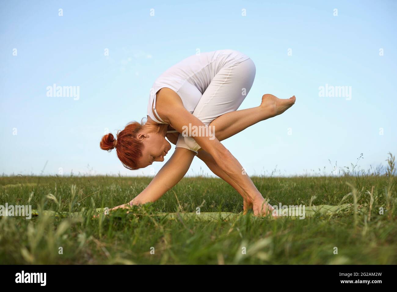 Junge Frau mit flexiblem Körper übt Yoga-Übungen auf dem Gras gegen den Himmel. Stockfoto