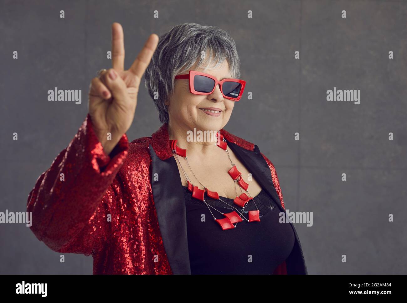 Glückliche ältere Frau in Pailletten-Party-Outfit und coole Brille tun Sieg Geste Stockfoto