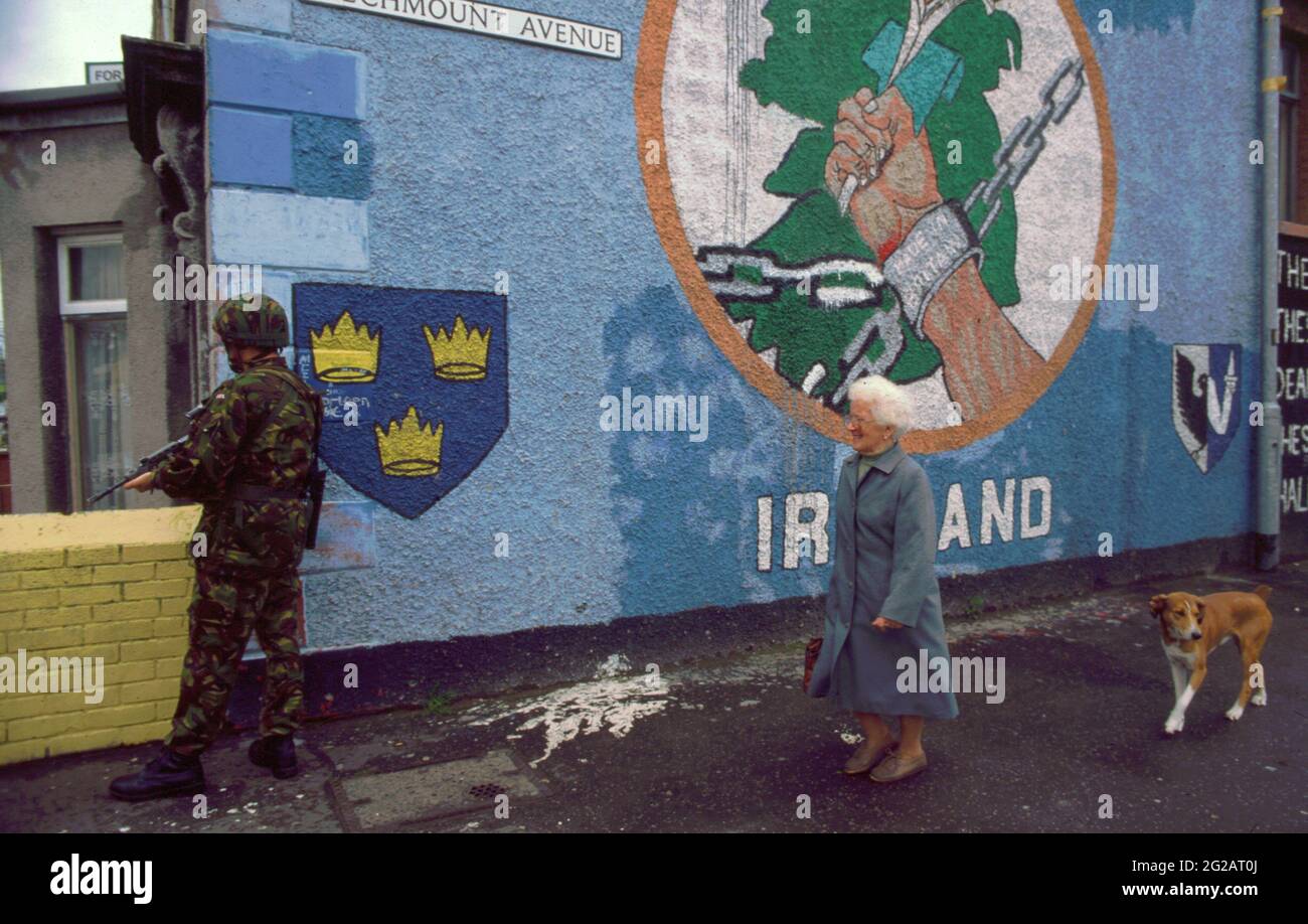 Belfast, Nordirland. Oktober 1993. Ein Soldat der britischen Royal Marines auf einer Patrouille auf der Beechmount Avenue und der Falls Road Kreuzung, als eine ältere Frau und ein Hund an einem republikanischen Wandgemälde für ein freies Irland vorbeikommen. Die Beechmount Avenue wurde als RPG Avenue bekannt, nachdem von diesem Standort aus eine mit Raketen angetriebene Granate auf britische Streitkräfte abgefeuert wurde. Stockfoto