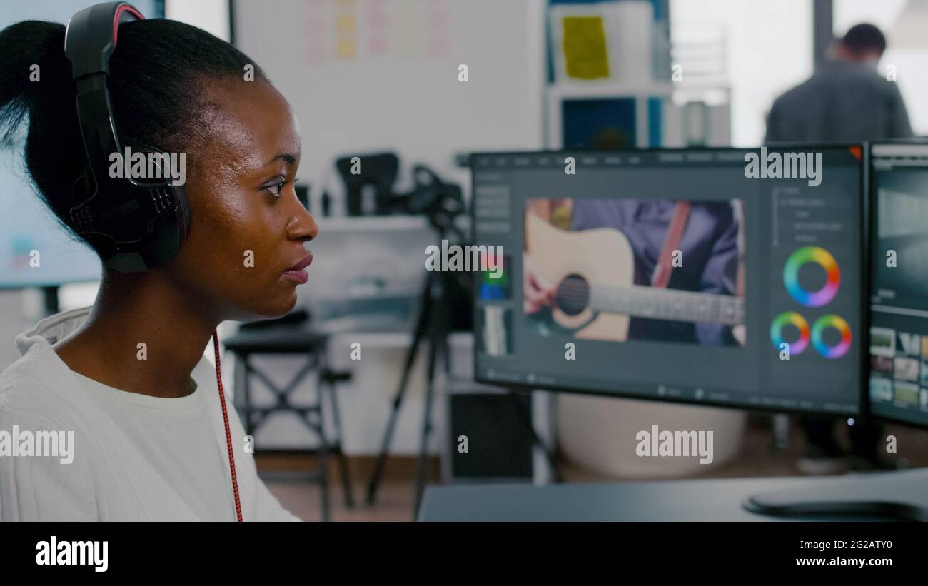 Schöne afrikanische Videofilmerin mit Kopfhörern, die Videomaterial auf dem pc bearbeitet und zwei Displays in einer Kreativagentur verwendet. Mitarbeiter, der Postproduktionssoftware verwendet, arbeitet im Büro einer Kreativagentur. Stockfoto