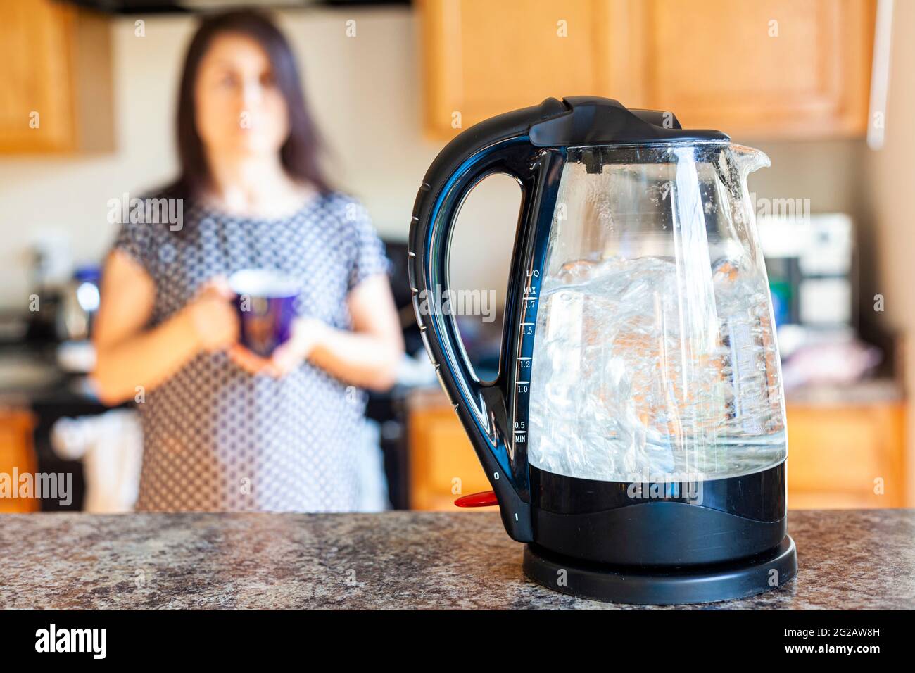 Eine kaukasische Frau wartet mit einer Tasse in den Händen, während Wasser in einem Glaskessel auf der Arbeitsplatte einer Küche kocht. Im Wasserkocher gibt es Mess Stockfoto