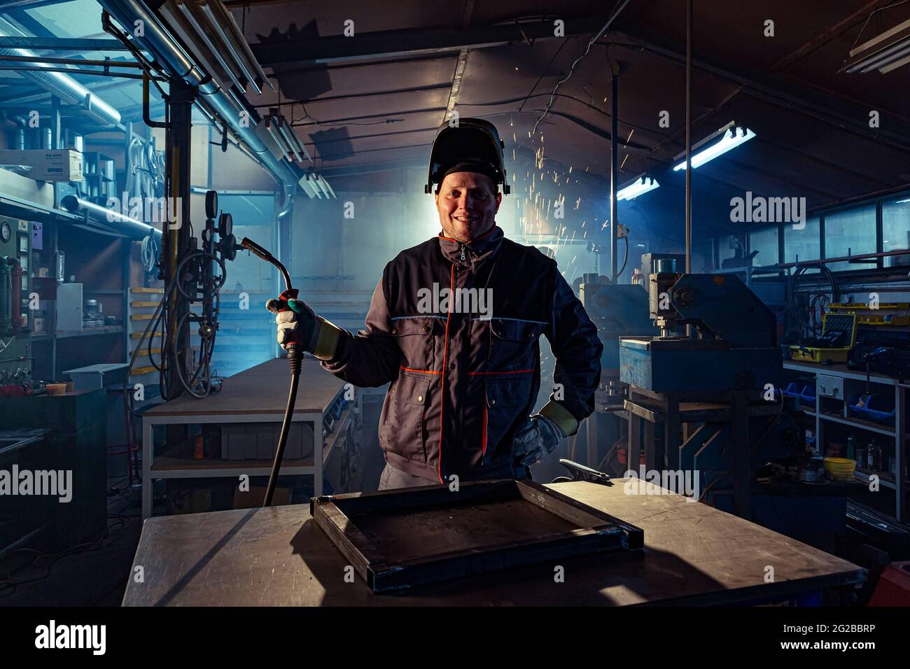 Porträt eines lächelnden Schweißers, der an seinem Arbeitsplatz in der Fabrik posiert, und es ist offensichtlich, dass der Meister mit dem Schweißen beschäftigt ist. Stockfoto