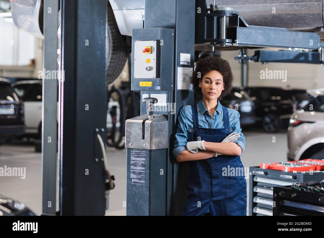 Junger afroamerikanischer Mechaniker, der mit gekreuzten Armen in der Nähe des angehobenen Autos steht und in der Garage auf die Kamera schaut Stockfoto