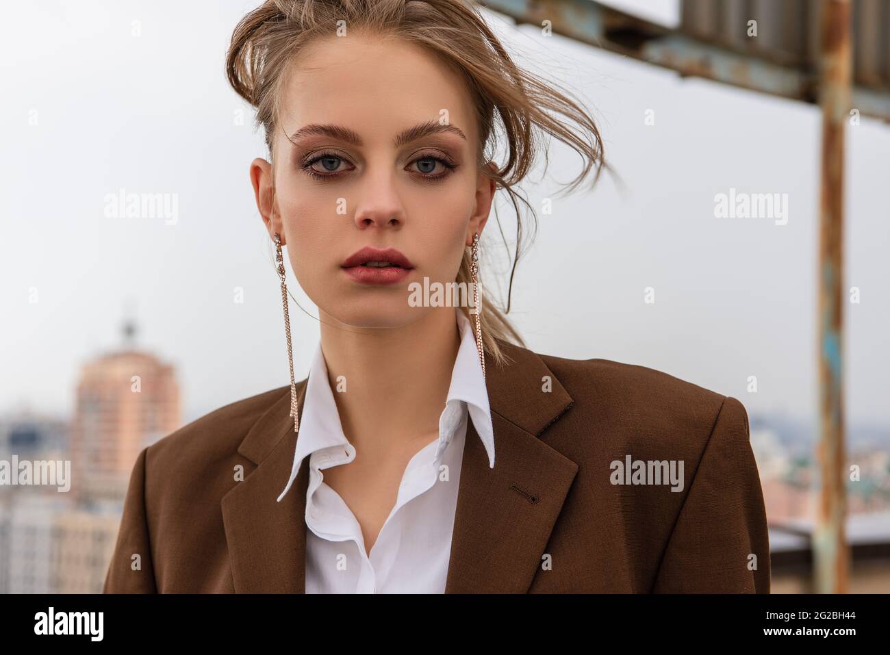 Junges Model in Ohrringen, weißem Hemd und brauner Jacke, das auf dem Dach die Kamera anschaut Stockfoto
