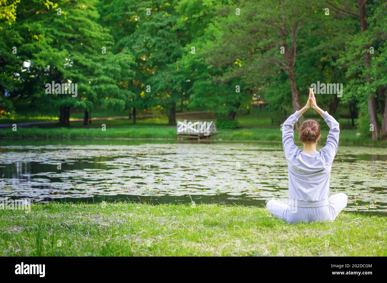 Junge Frau in Lotusposition, die am frühen Morgen auf grünem Gras sitzt. Yoga im Park mit Blick auf den See, mit Sonnenlicht. Ruhe und Meditation. Stockfoto