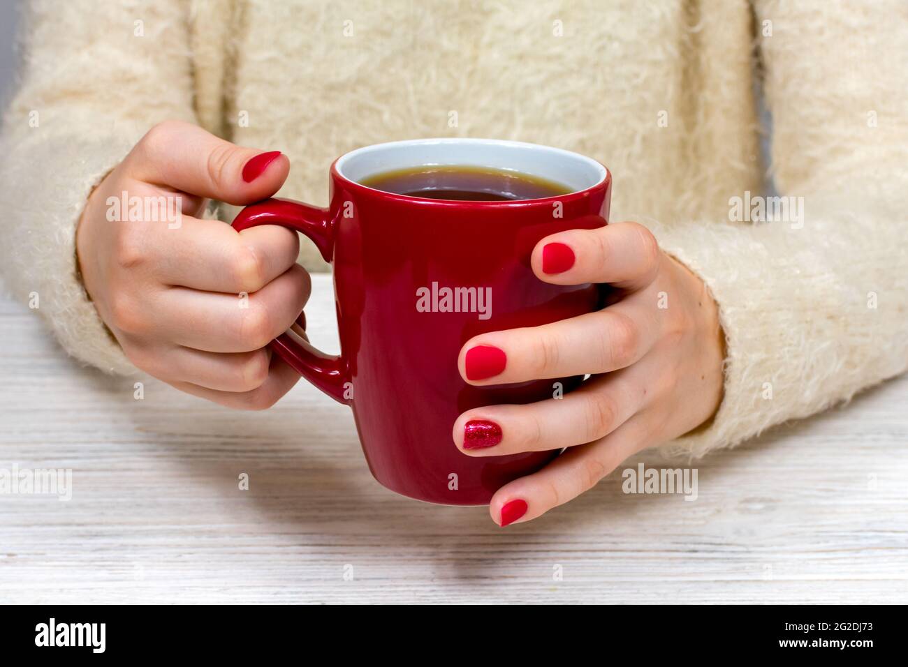 Weibliche Hände halten eine rote Tasse vor dem Hintergrund eines weißen flauschigen Pullovers mit einer schönen roten Winter-Maniküre. Getränk, Mode, Morgen. Neujahr Stockfoto