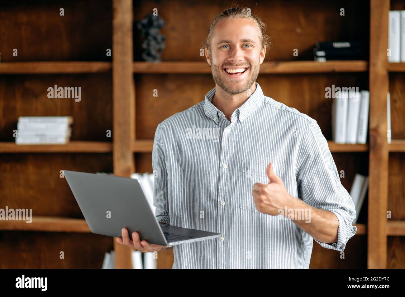 Ein glücklicher, gutaussehender junger, erwachsener amerikaner steht in einem stylischen Hemd im Büro, hält einen Laptop in der Hand, zeigt Daumen nach oben, blickt auf die Kamera und lächelt freundlich Stockfoto