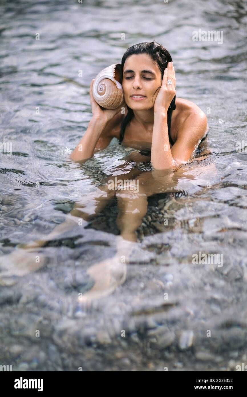 Weibchen im Wasser hält die Muschel am Ohr und genießt das Rauschen der Wellen. Sommerurlaub und Entspannungskonzept. Stockfoto