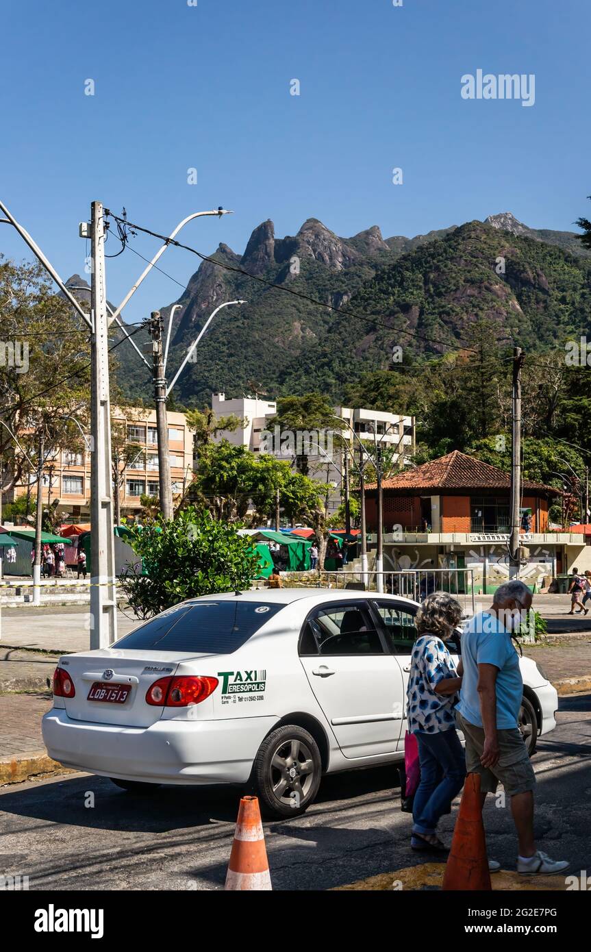 Ein weißes Taxi, das an den Straßen des Higino da Silveira-Platzes vorbeifährt, mit Teilblick auf die Zelte, Gebäude und einen großen Berghang hinten. Stockfoto