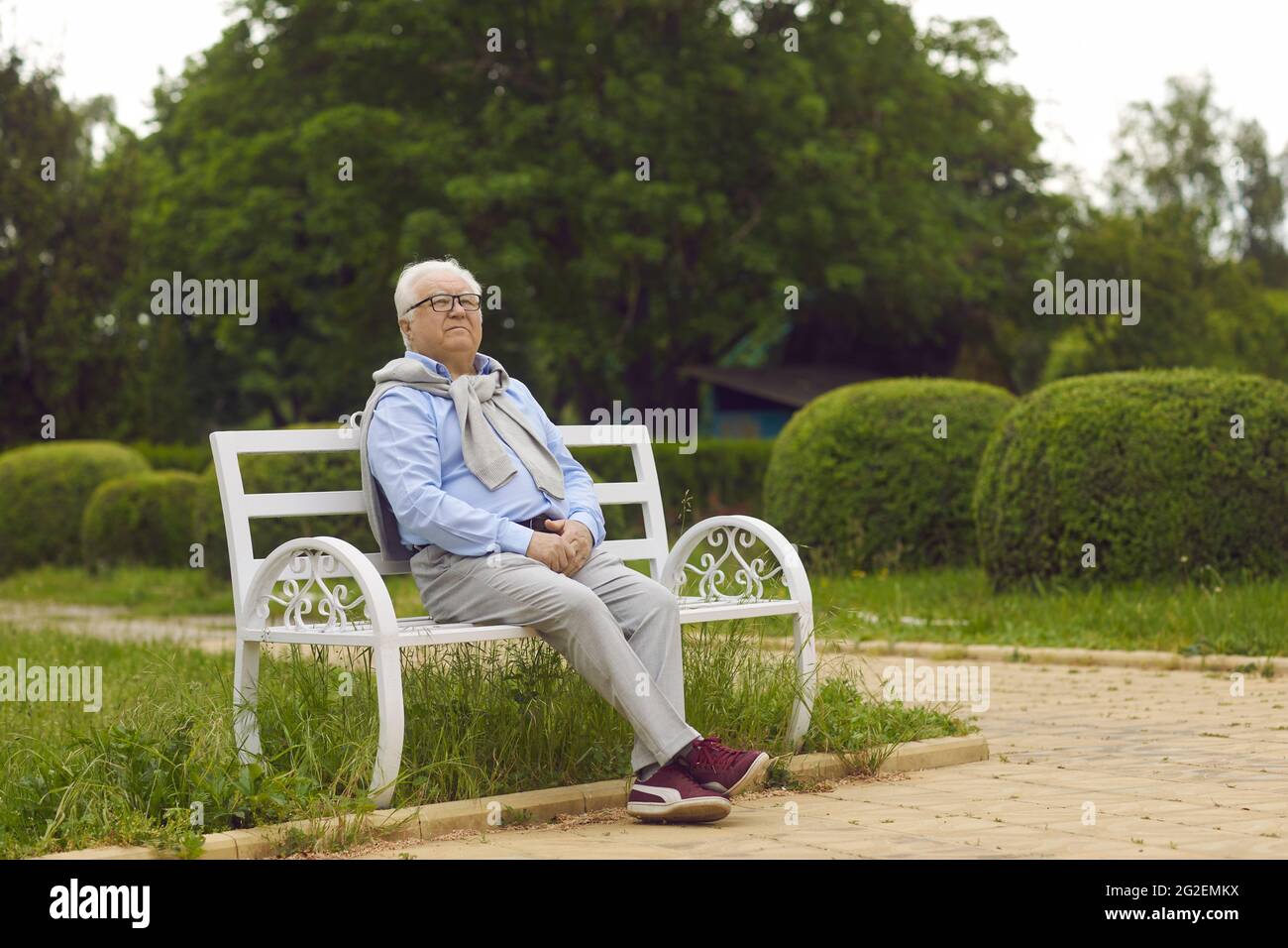 Älterer Mann bewundert die Natur, während er auf einer weißen Bank in einem grünen Sommerpark sitzt Stockfoto