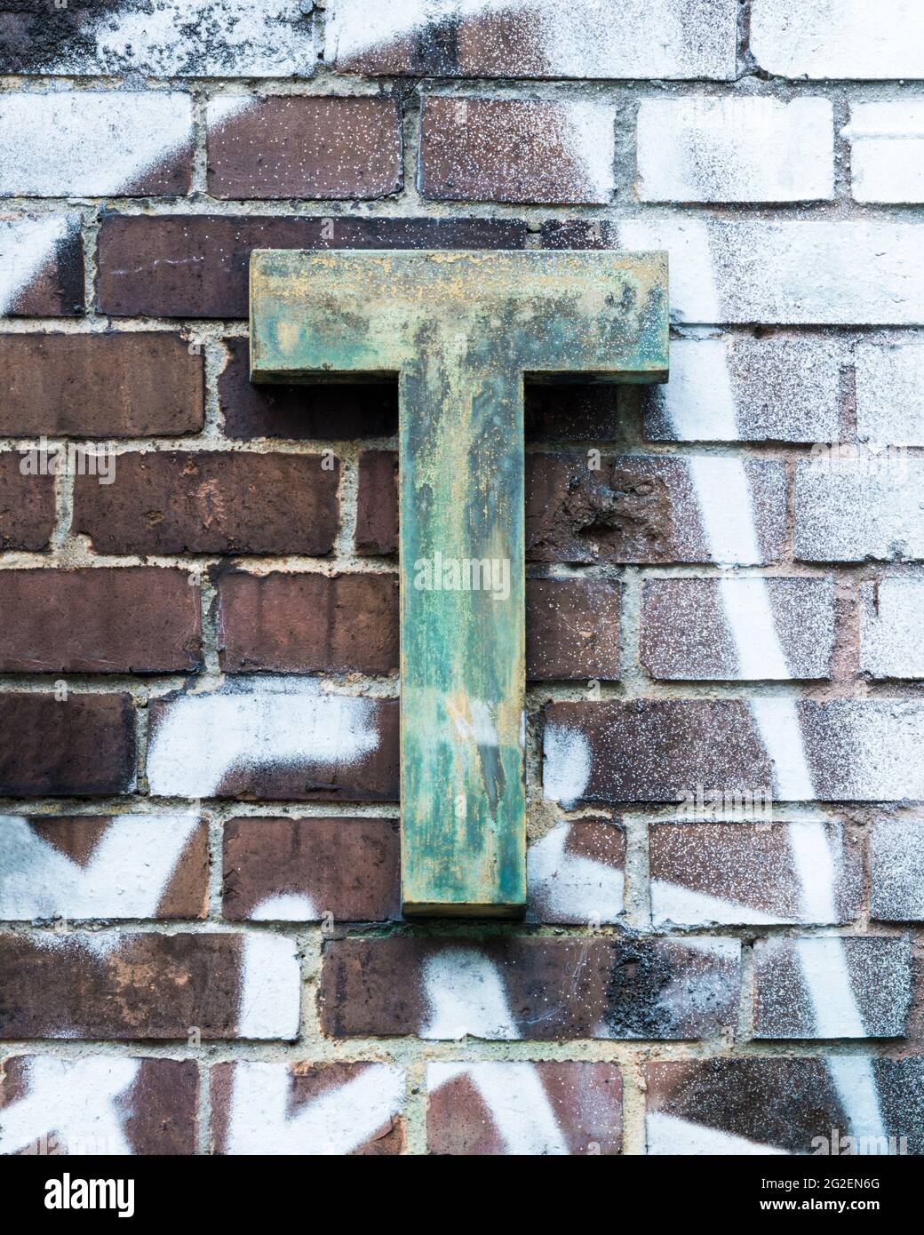 Verwitterter Metallbuchstabe T vor einer mit weißer Sprühfarbe überdeckten Ziegelwand Stockfoto