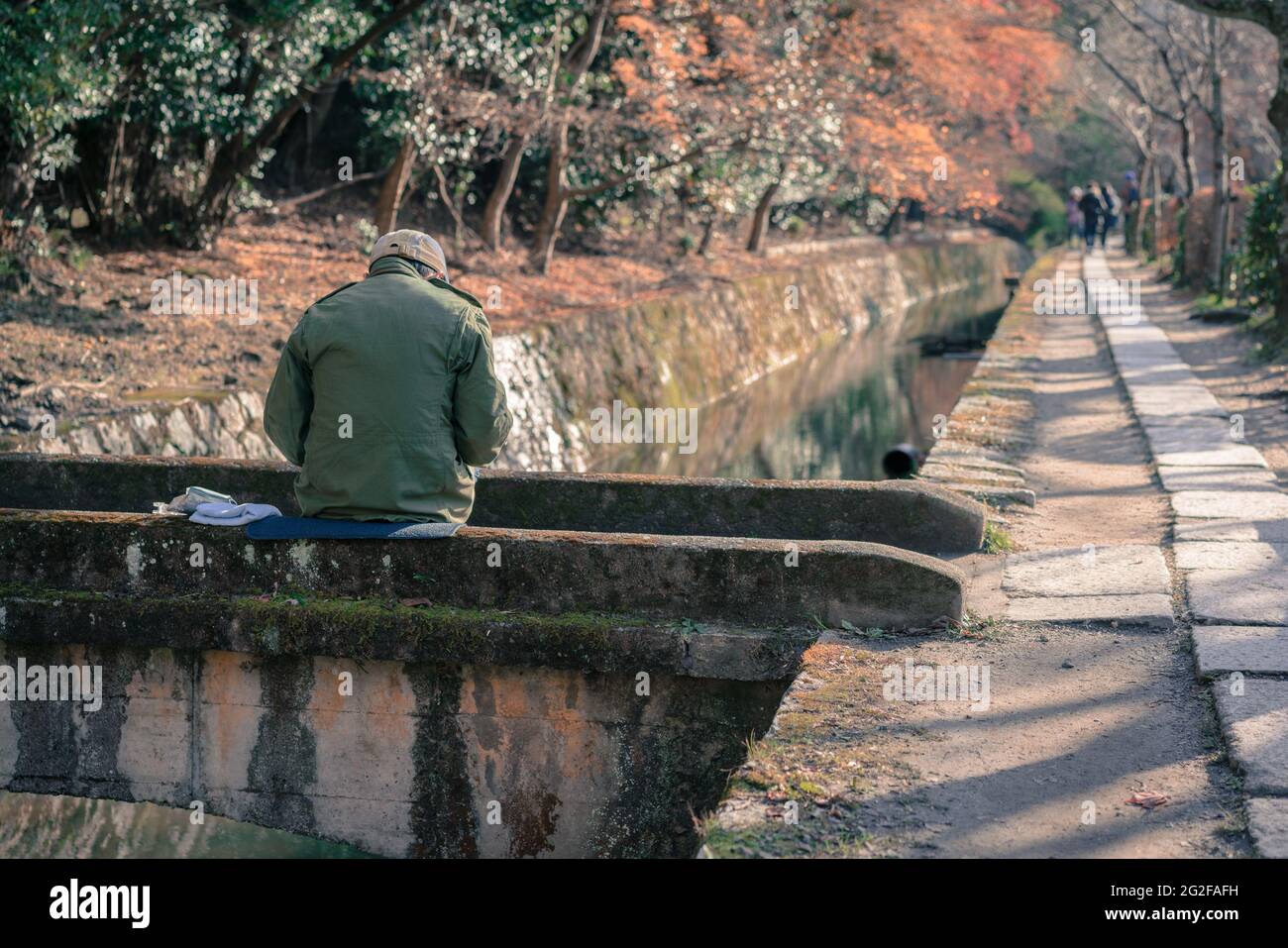 Ein älterer Mann, der auf einer Steinbrücke über einen Wasserkanal sitzt, malte das Bild des Philosophen-Spaziergangs in Kyoto, Japan. Herbstfarben im historischen Japan Stockfoto