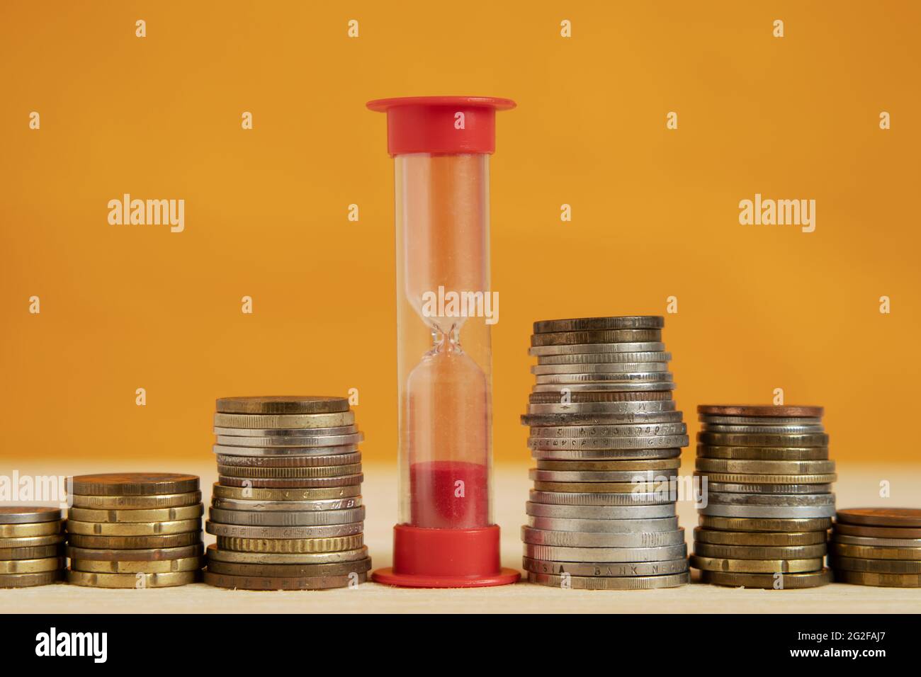 Eine rote Sanduhr steht in einer Reihe mit Münzstapeln. Das Konzept von Zeit und Geld. Nahaufnahme. Orangefarbener Hintergrund Stockfoto