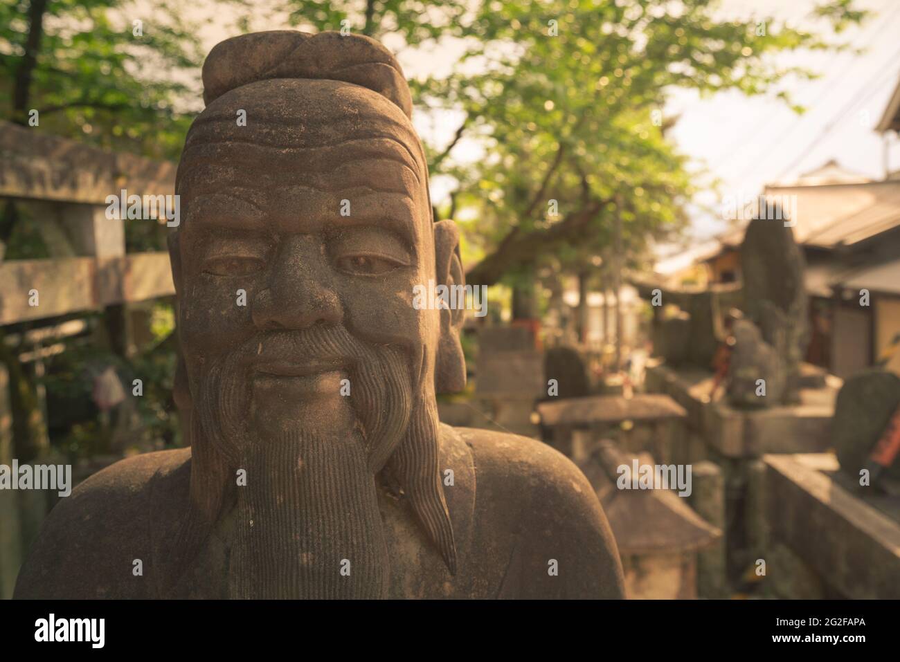 Kopfaufnahme einer Steinstatue eines weisen Mannes im asiatischen Stil. Statue auf dem buddhistischen Friedhof im Komplex des Fushimi-Inari-Schreins in Kyoto, Japan. Goldene Stunde Stockfoto