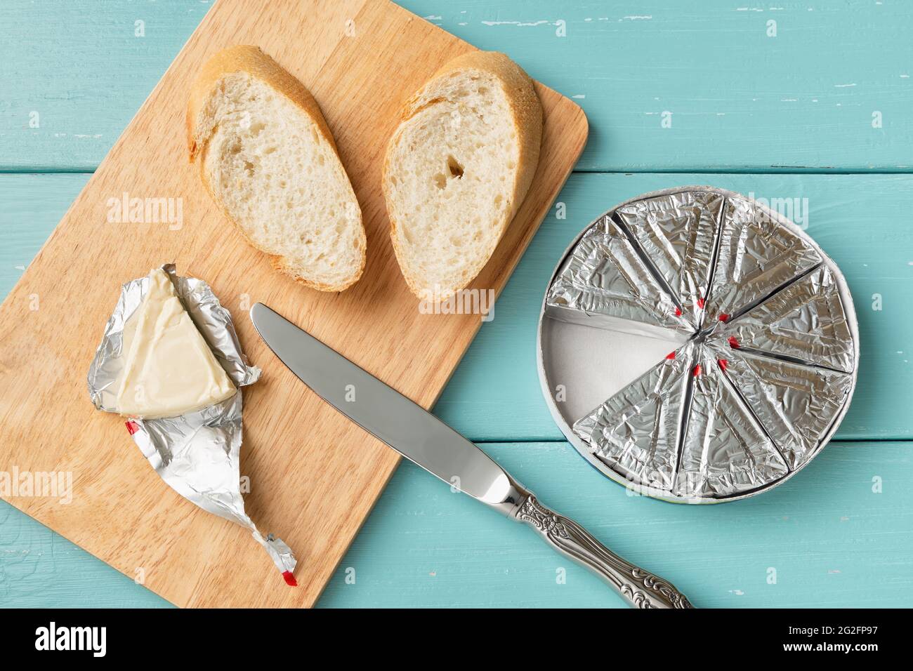 Sandwich mit verarbeitetem Frischkäse und geschnittenem Brot auf einem Schneidebrett über einem blauen Holztisch zubereiten. Kleine dreieckige Käsestücke in Folienverpackung Stockfoto