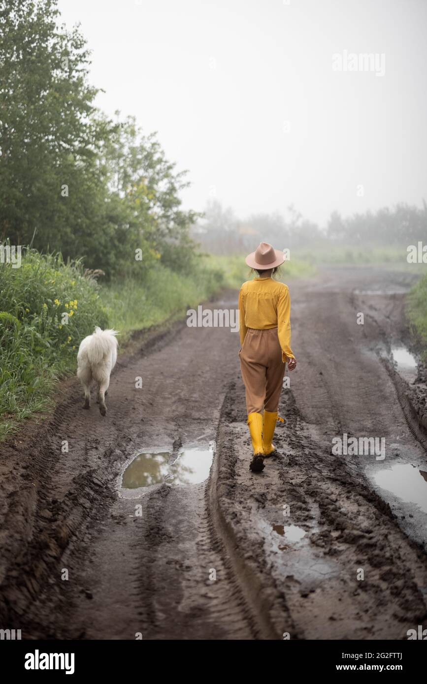 Bäuerin in gelben Gummistiefeln, die nach dem Regen auf einer schlammigen Straße mit Schafhund unterwegs ist. Stockfoto