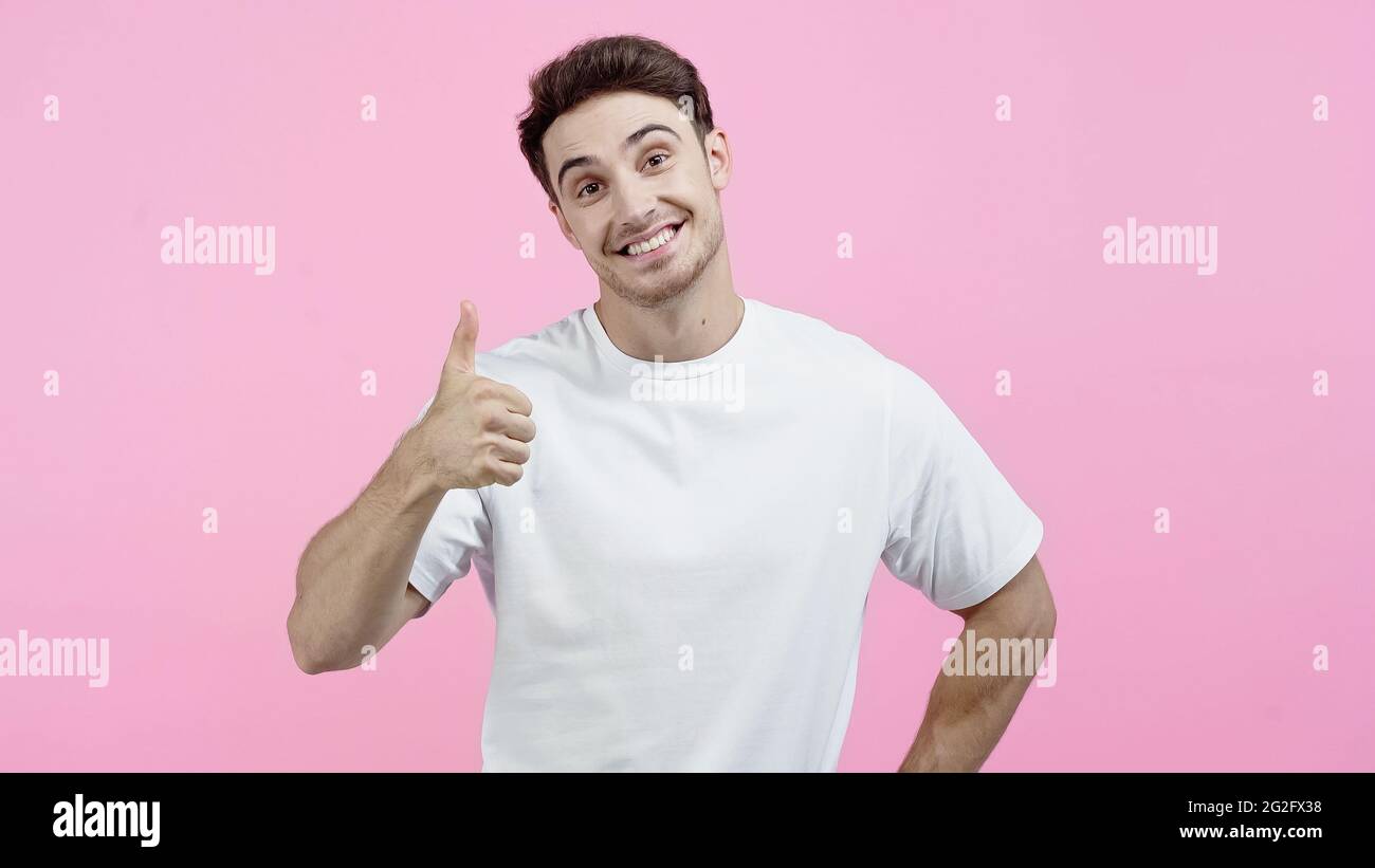 Lächelnder Mann in weißem T-Shirt, der die Kamera anschaut und den Daumen isoliert auf Pink zeigt Stockfoto