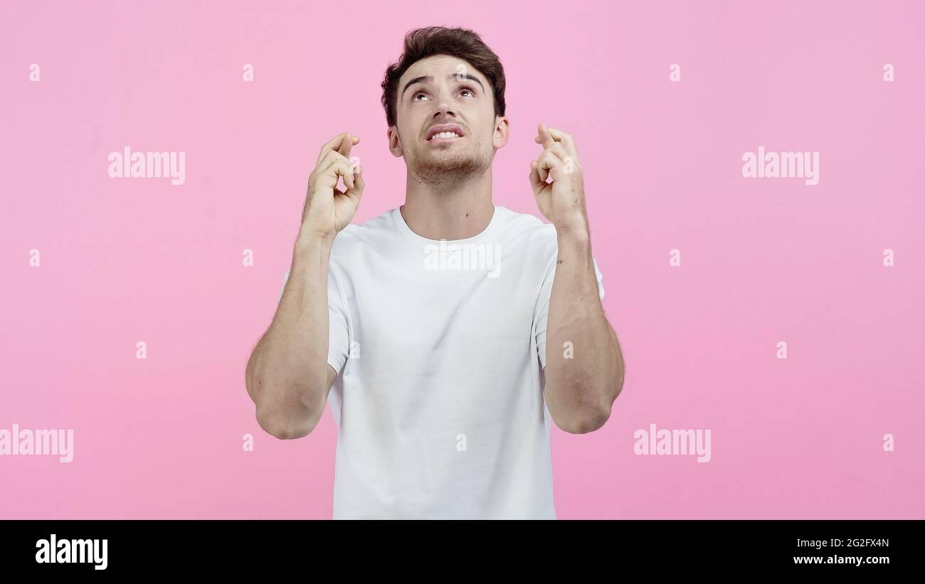 Junger Mann im weißen T-Shirt, der gekreuzte Daumen zeigt und isoliert auf Pink aufschaut Stockfoto