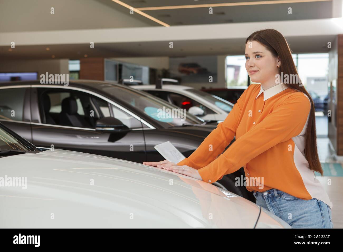 Schöne glückliche junge Frau, die träumerisch wegschaut, während sie sich für ein neues Auto im Autohaus entscheidet Stockfoto