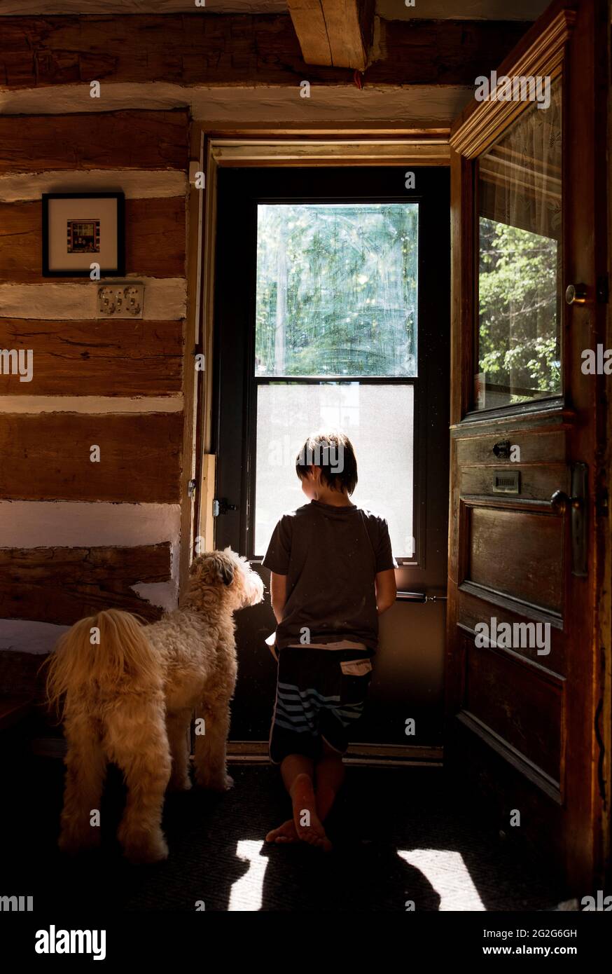 Junge und sein Hund blicken an sonnigen Tagen aus der Tür eines Blockhütten-Hauses. Stockfoto
