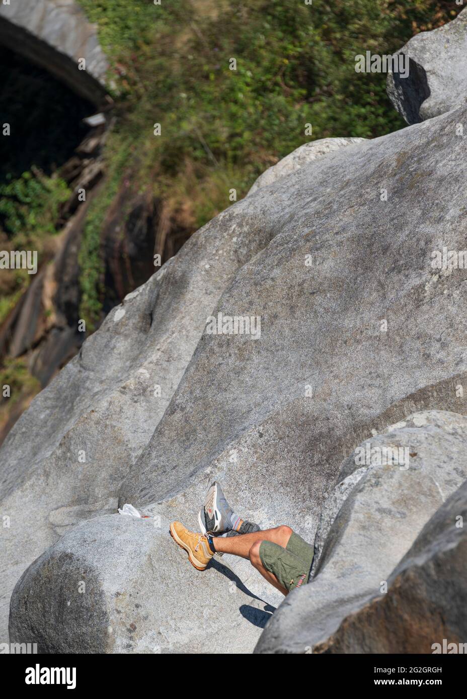 Impressionen von Lavertezzo im Verzasca-Tal, Bezirk Locarno, Kanton Tessin in der Schweiz: Beliebtes Ausflugsziel zum Wandern, Flusstauchen und Schwimmen. Zwei Männer entspannen sich in der Sonne auf den Granitfelsen unter der Brücke, nur ihre Beine sind sichtbar. Stockfoto