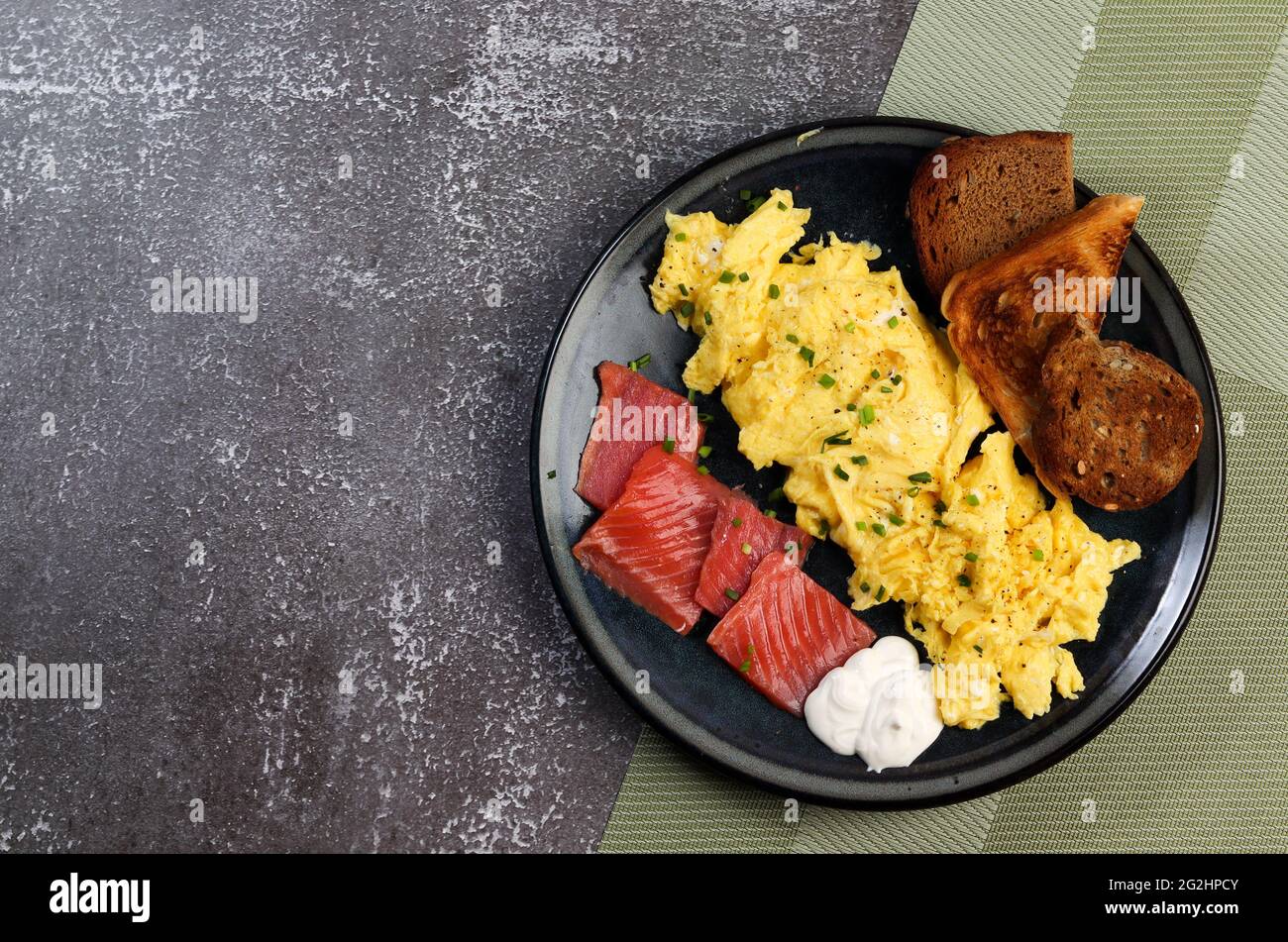 Rührei mit Lachs und Toast auf einem runden Teller auf dunklem Grund. Draufsicht, flach liegend Stockfoto