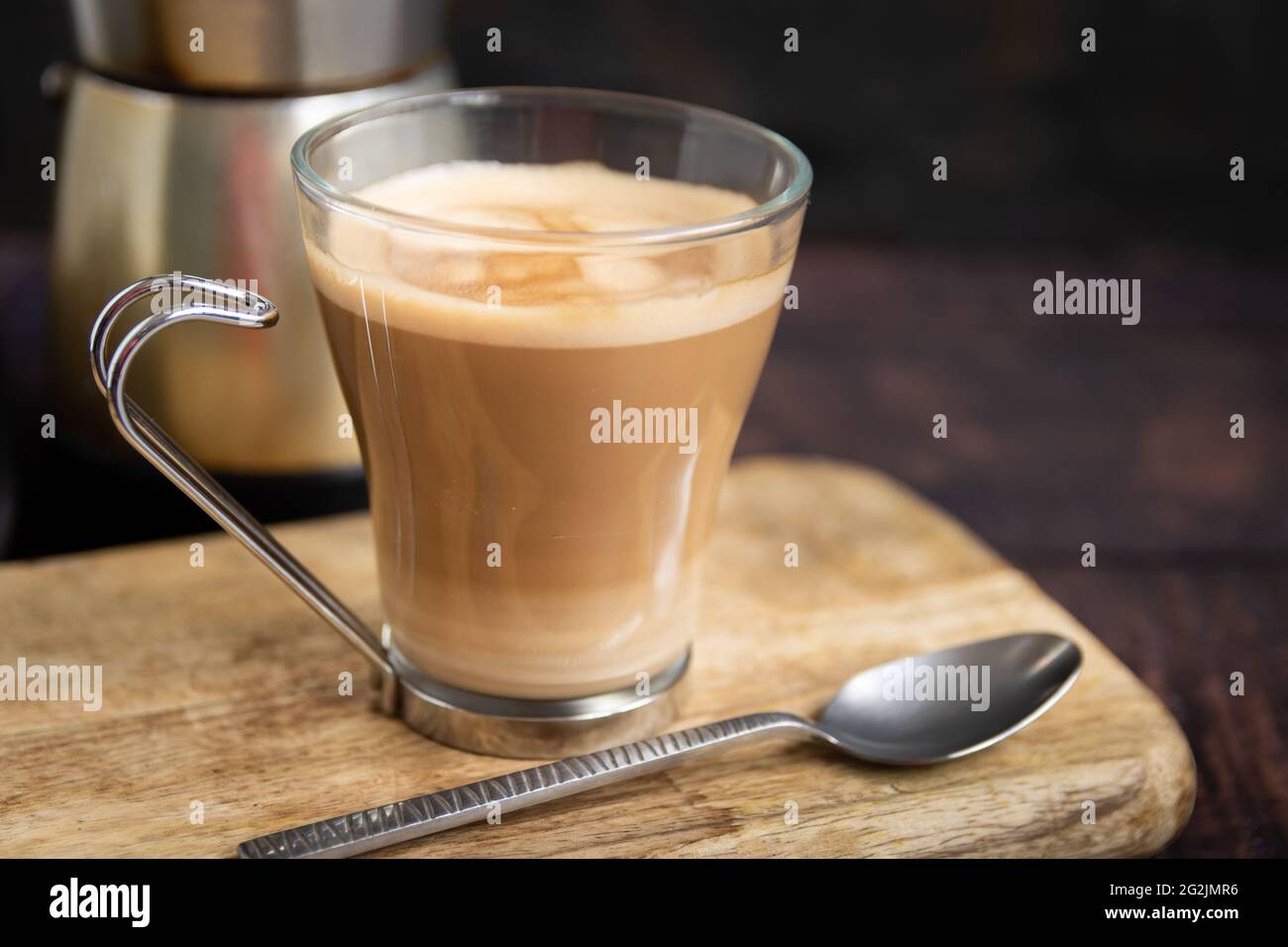 Tasse Kaffee mit Milch und Teelöffel auf Holztisch und italienische Kaffeekocher im Hintergrund Stockfoto