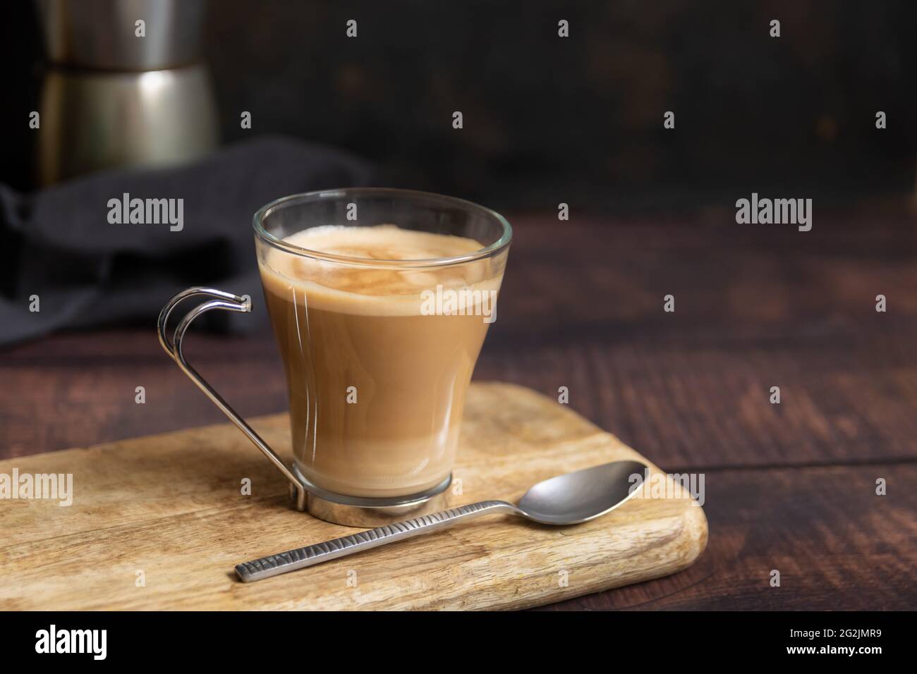 Tasse Kaffee mit Milch und Teelöffel auf Holztisch und italienische Kaffeekocher im Hintergrund Stockfoto