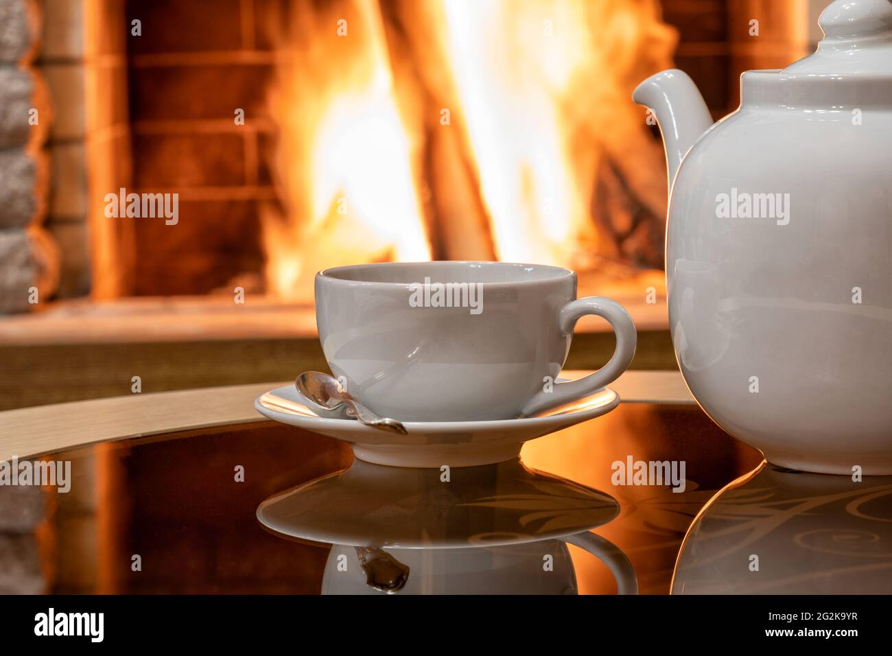 Gemütliche Kamin-Szene und weiße Tasse Tee und Teekannen auf einem Tisch, Spiegelung von einem Feuer auf einem Glastisch. Stockfoto