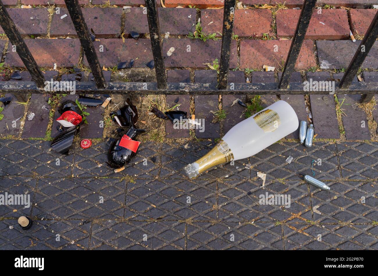 Zerbrochene Bierflasche, Champaign-Flasche und Lachgaskanister aus Metall mit Lachgas aus Lachgas auf dem Bürgersteig neben einem Metallzaun. London - 12. Juni 2021 Stockfoto