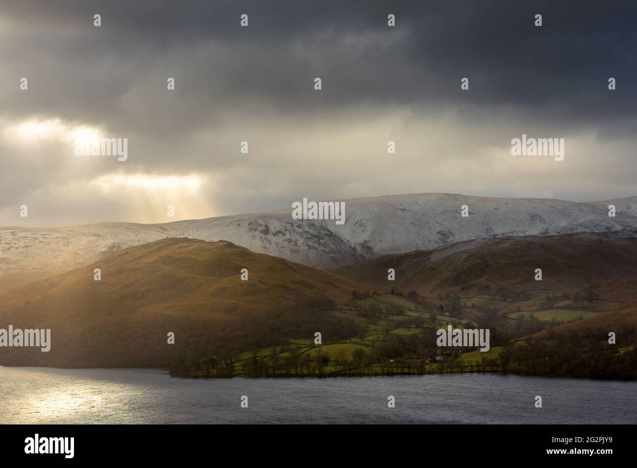 Dramatische Lichtstrahlen brechen durch dunkle Wolken über schneebedeckten Cumbrian Bergen, die Ullswater im Lake District umgeben. Stockfoto
