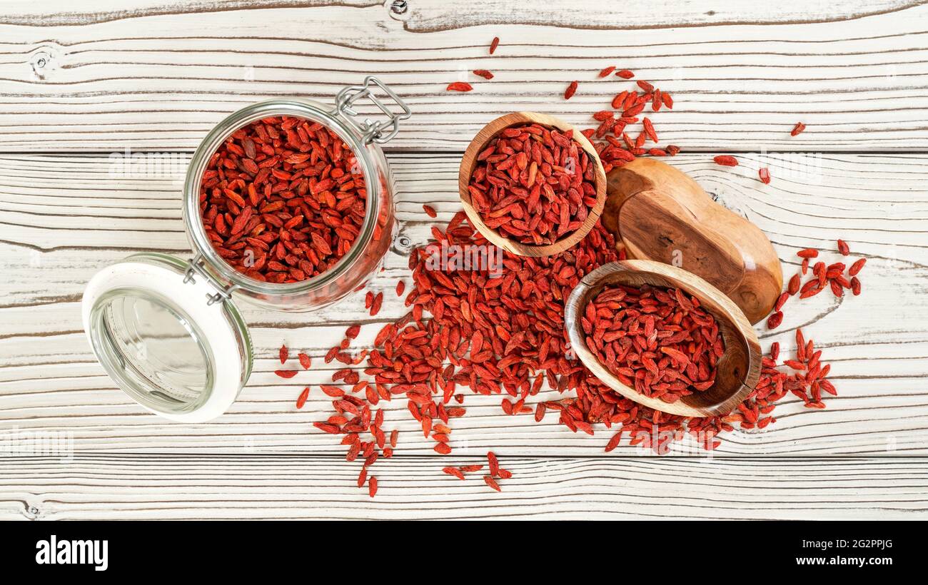 Getrocknete Goji aka. wolfberry Samen in Glasflasche, einige in Holzschüsseln und verschüttet auf weißen Brettern Schreibtisch, Blick von oben Stockfoto