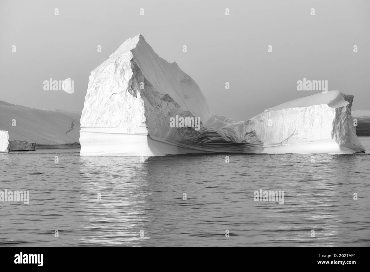 Schwarz-Weiß-Foto eines riesigen schwimmenden Eisbergs in der Dämmerung mit dem Mond auf der linken Seite. Scoresby Sund, Kangertittivaq, Grönland, Dänemark Stockfoto