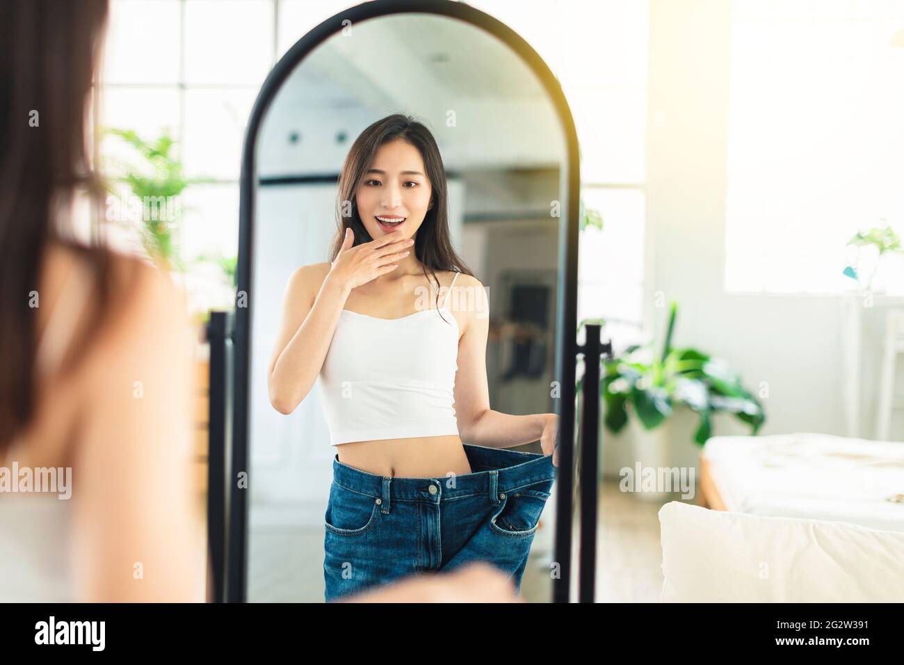 Die junge Frau, die große Jeans trägt, schaut sich ihre Figur vor dem Spiegel an, sehr glücklich, dass es ihr gelungen ist, Gewicht zu verlieren. Stockfoto