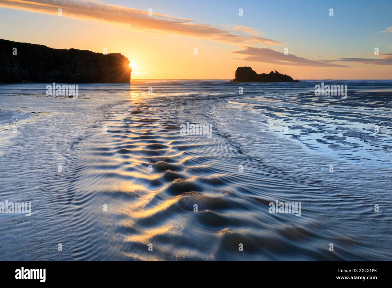 Sandkräuseln und ein Fluss führen das Auge des Betrachters in Richtung Chapel Rock am Perranporth Beach in Cornwall. Das Bild wurde kurz vor Sonnenuntergang aufgenommen. Stockfoto