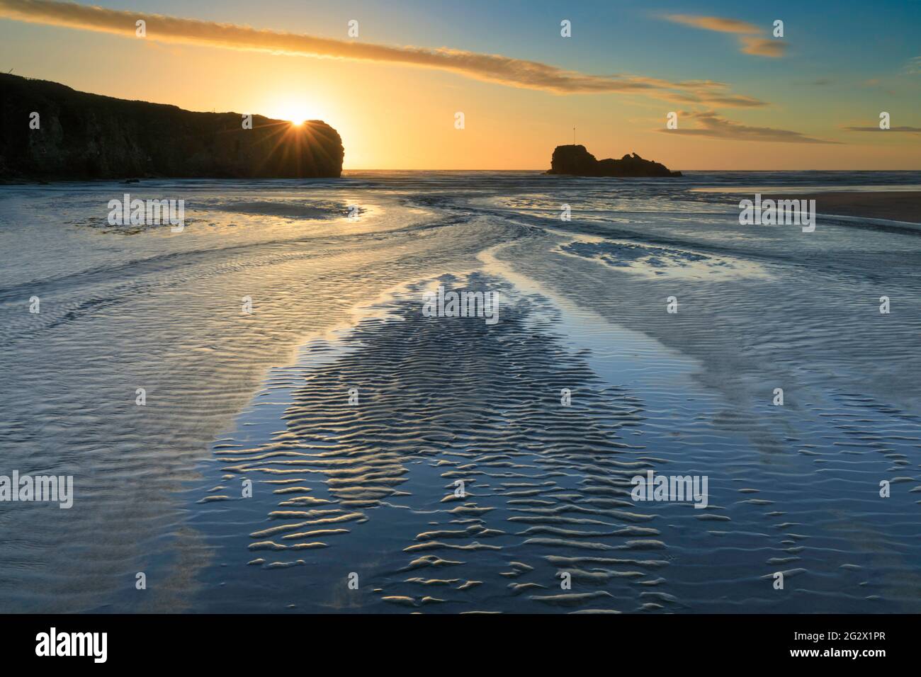 Sandkräuseln und ein Fluss führen das Auge des Betrachters in Richtung Chapel Rock am Perranporth Beach in Cornwall. Das Bild wurde kurz vor Sonnenuntergang aufgenommen Stockfoto