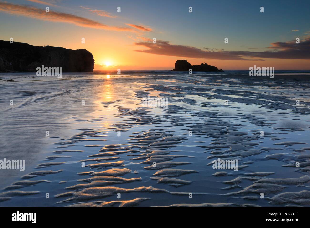 Sandkräuseln und ein Fluss führen das Auge des Betrachters in Richtung Chapel Rock am Perranporth Beach in Cornwall. Das Bild wurde kurz vor Sonnenuntergang aufgenommen Stockfoto